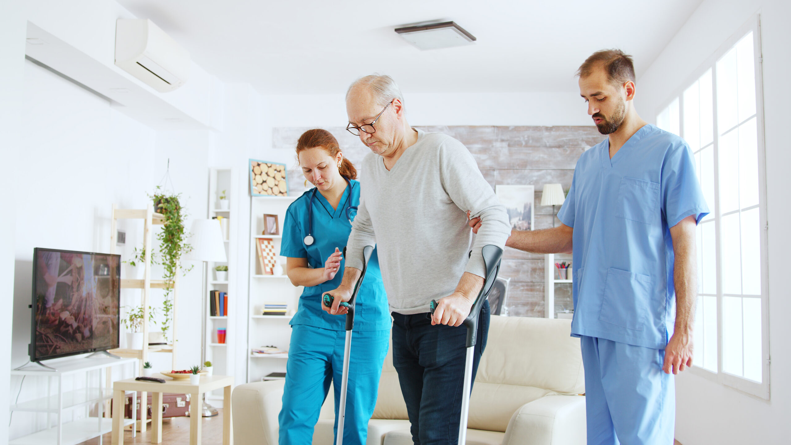 http://Team%20of%20nurses%20or%20social%20workers%20helping%20an%20old%20disabled%20man%20to%20walk%20with%20his%20crutches%20out%20of%20the%20nursing%20home%20room